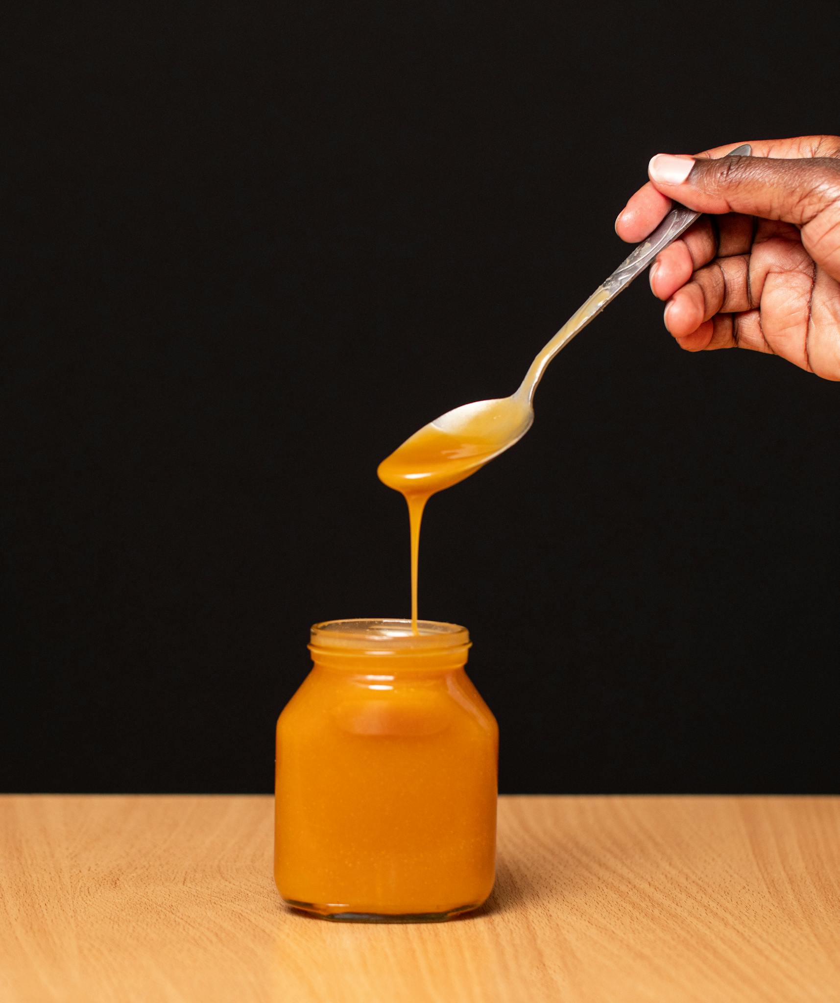 Close-up of honey dripping from a spoon into a glass jar against a dark background.